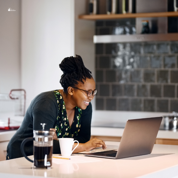 A stay-at-home mom smiles while working on a laptop in her kitchen, with a cup of coffee beside her.