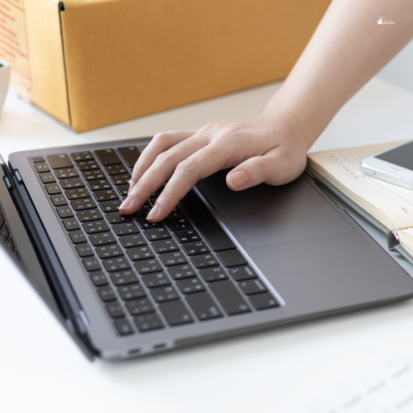 A close-up of a hand typing on a laptop keyboard, with a cardboard box and notebook in the background.