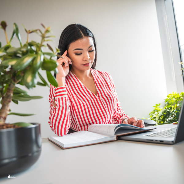 A woman in a striped blouse sits at a desk, thoughtfully reading a book next to her open laptop.