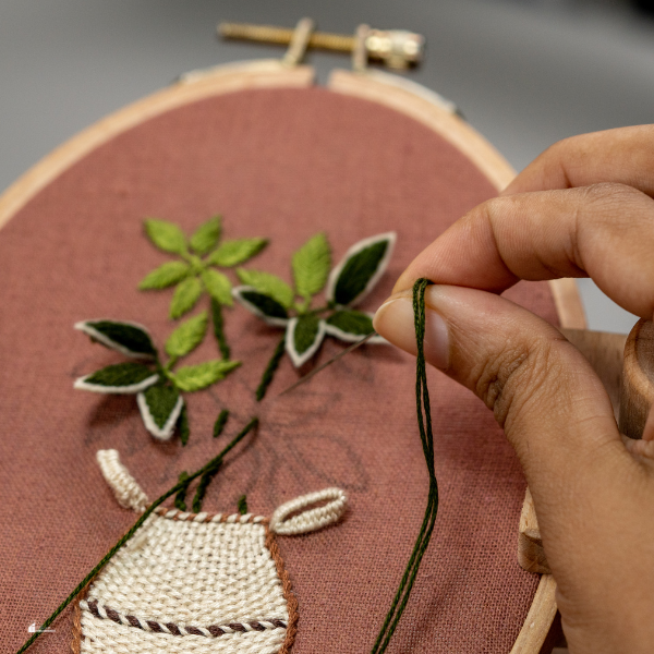 A close-up of a hand working on embroidery, creating a detailed floral design on a fabric hoop.