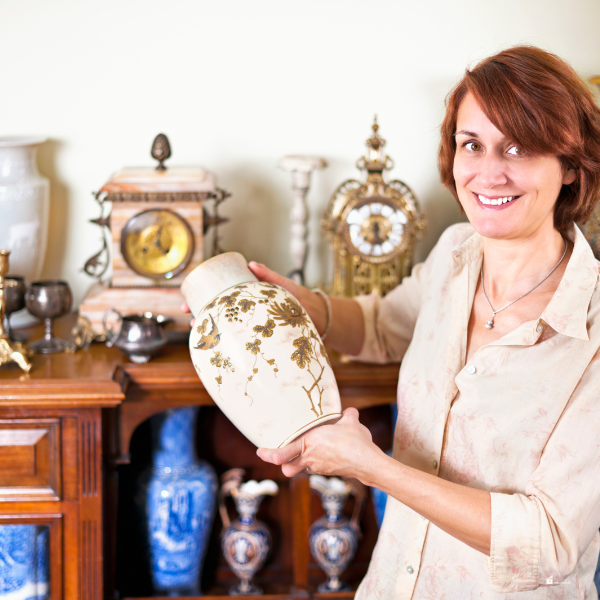 A woman smiles while holding a vintage vase, surrounded by various collectibles.