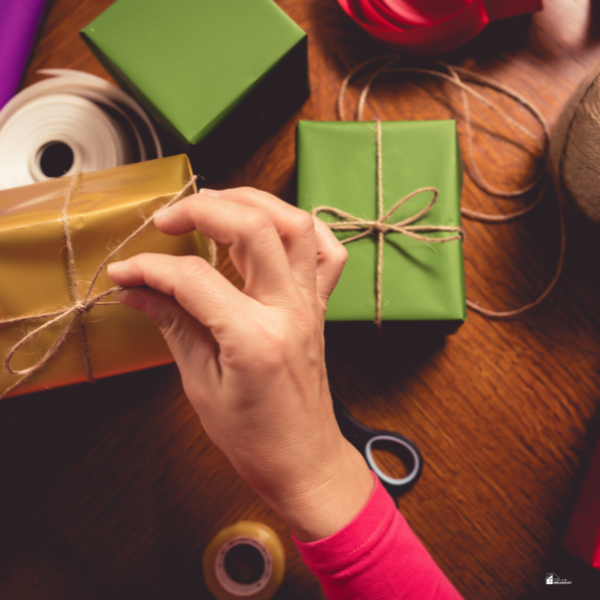 A close-up of hands tying a bow with twine on a gift box, surrounded by other wrapped presents on a wooden table.