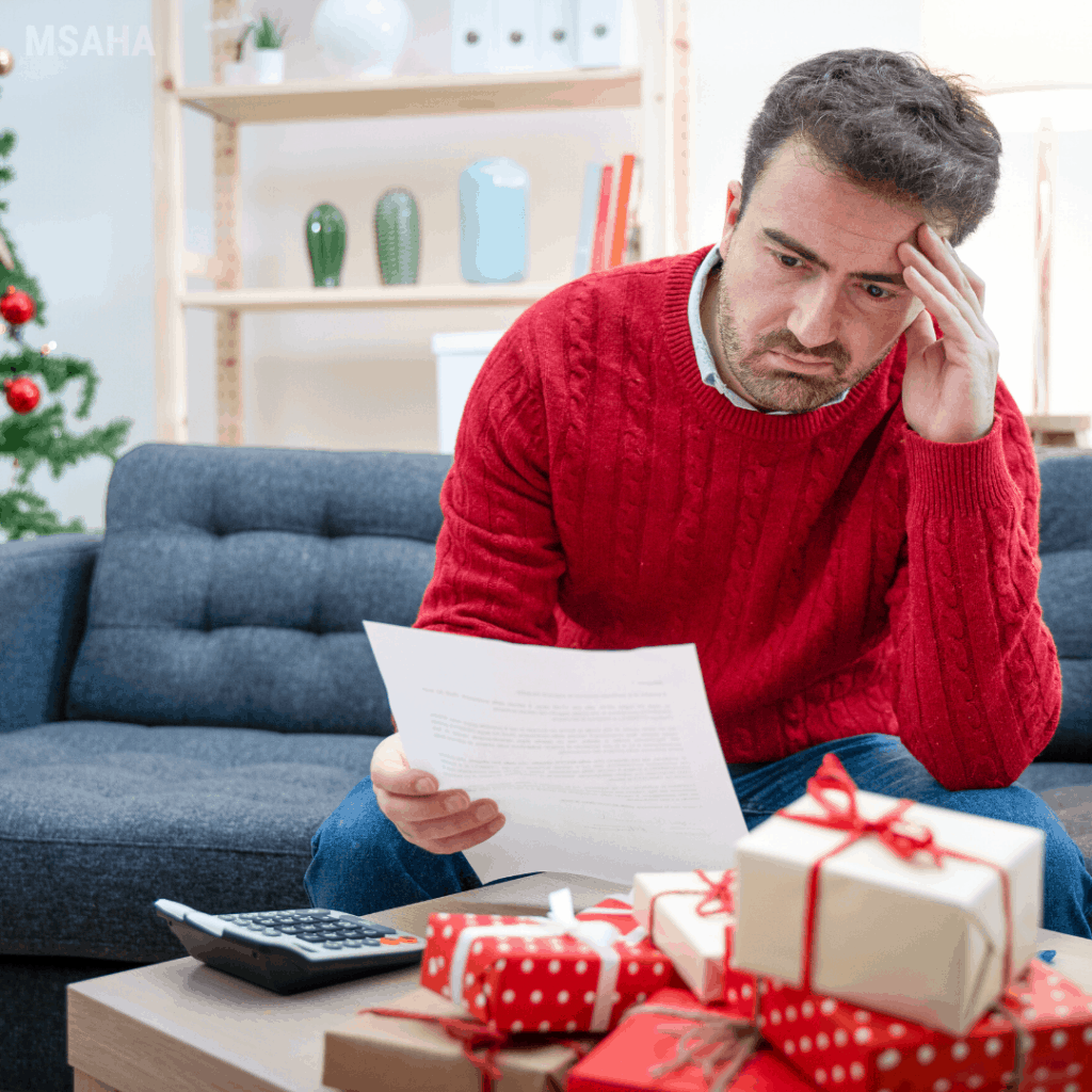 A man in a red sweater sits by a Christmas tree, looking stressed while reviewing bills or a budget with wrapped gifts in front of him, symbolizing holiday financial concerns and overspending.