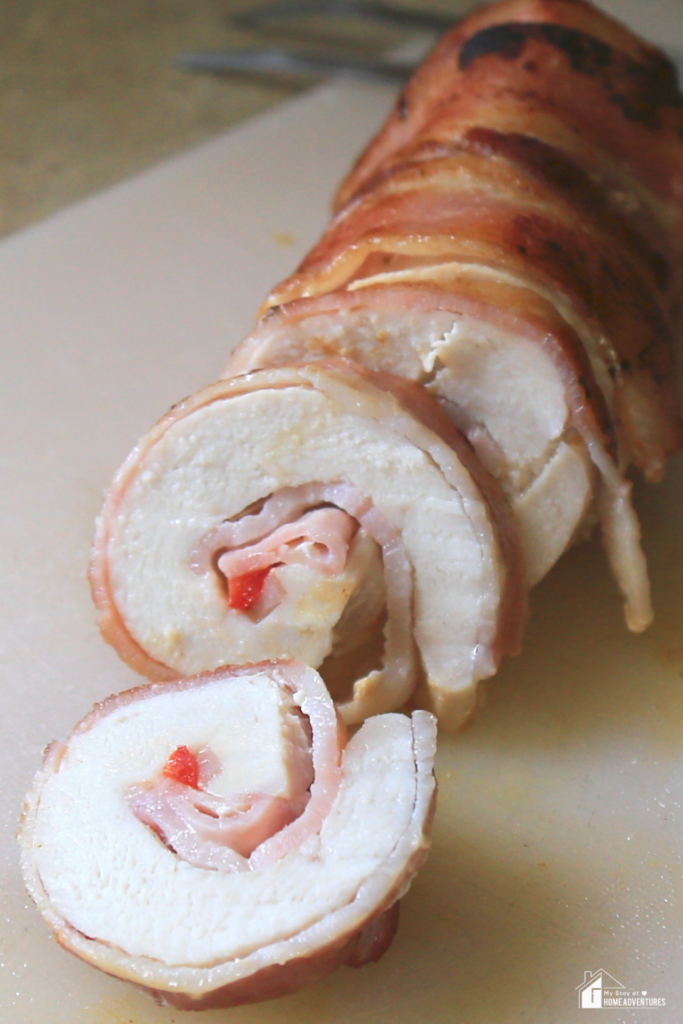 A close-up of sliced bacon-wrapped chicken roll-ups stuffed with ham and red peppers, displayed on a cutting board.