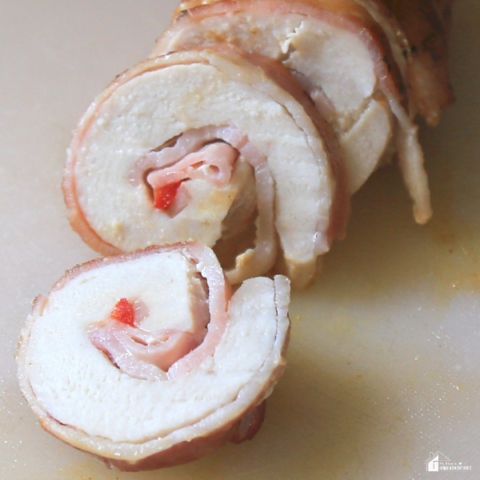 A close-up of sliced bacon-wrapped chicken roll-ups stuffed with ham and red peppers, displayed on a cutting board.