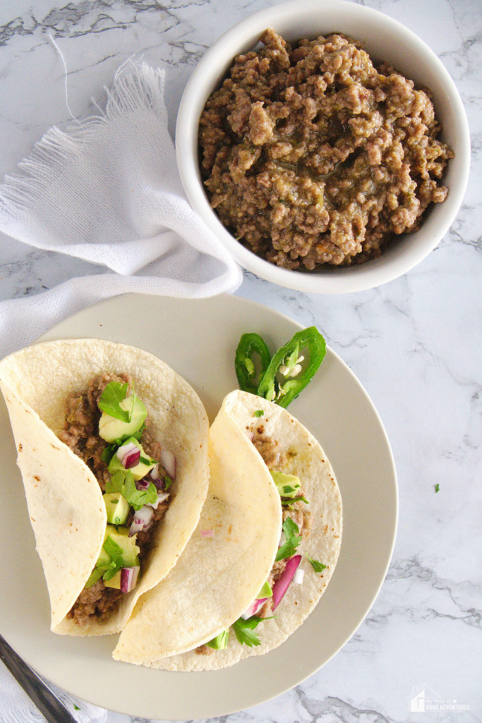 A bowl of Salsa Verde Beef Filling alongside two soft tacos filled with the beef mixture, garnished with fresh cilantro, red onion, and jalapeño slices.