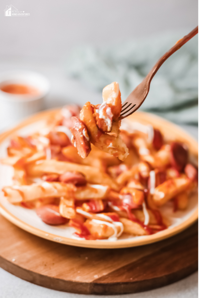 A fork lifting a bite of Salchipapas, a dish of crispy fries and sliced sausage, topped with ketchup and mayonnaise, served on a plate.