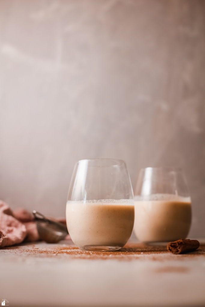 Two glasses of creamy Rompope, a traditional Mexican holiday drink, placed on a festive table with a cinnamon stick, perfect for Christmas or holiday gatherings.