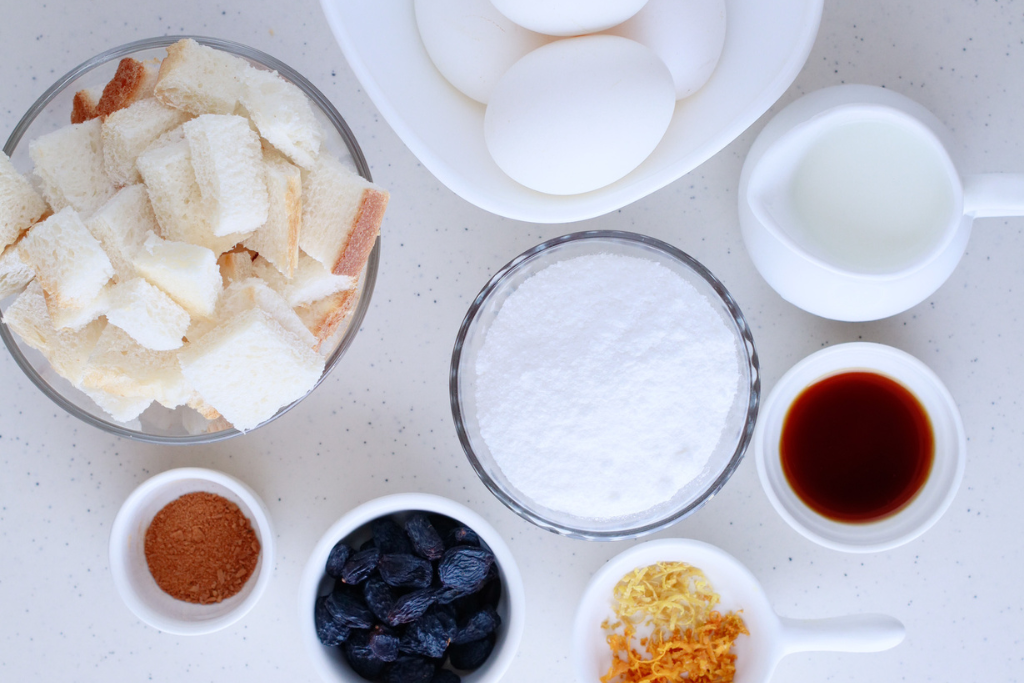 A top-down view of the key ingredients for Budín de Pan, a traditional Puerto Rican bread pudding, including cubed bread, eggs, milk, sugar, cinnamon, raisins, vanilla, and citrus zest.