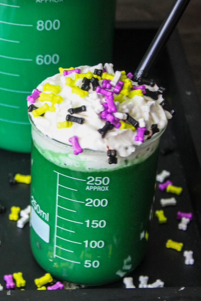 A close-up of a beaker filled with green Halloween hot cocoa, decorated with whipped cream and Halloween-themed bone sprinkles, ideal for monster-themed Halloween drinks.