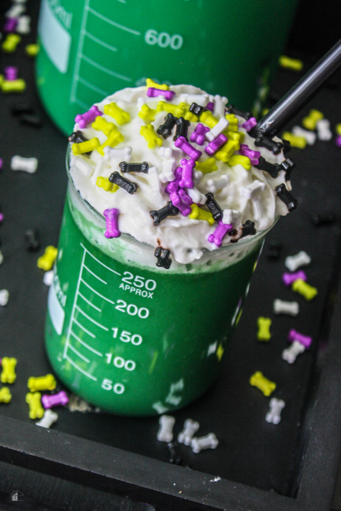 A close-up of a beaker filled with green Halloween hot cocoa, decorated with whipped cream and Halloween-themed bone sprinkles, ideal for monster-themed Halloween drinks.