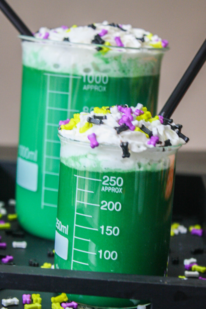 A vibrant green monster hot cocoa served in laboratory-style beakers, topped with whipped cream and spooky sprinkles, perfect for a fun and kid-friendly Halloween drink recipe.