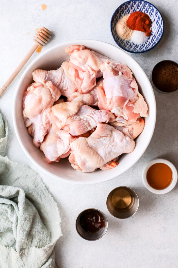 A top-down view of raw chicken wings in a white bowl, surrounded by spices, honey, bourbon, and other ingredients, ready to make crispy air fryer chicken wings with a sweet and smoky bourbon honey glaze.