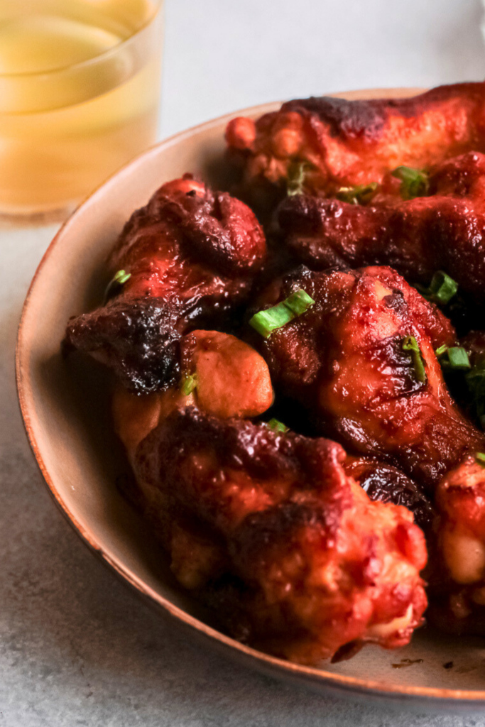 A close-up of crispy honey bourbon chicken wings coated in a sticky, caramelized glaze, garnished with green onions, served in a bowl with a glass of bourbon in the background, perfect for air fryer appetizer recipes and game day snacks.