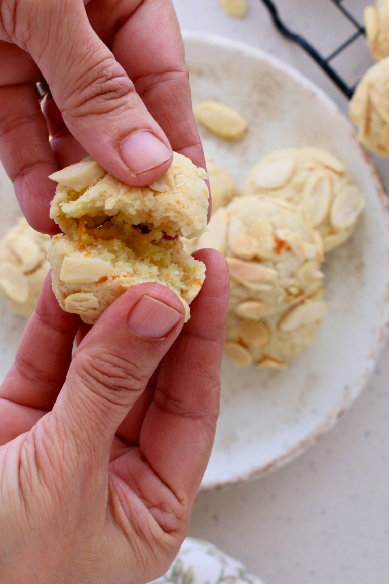 A close-up of hands breaking apart a Greek Almond Cookie, highlighting its soft, crumbly texture with visible orange zest and slivered almonds.