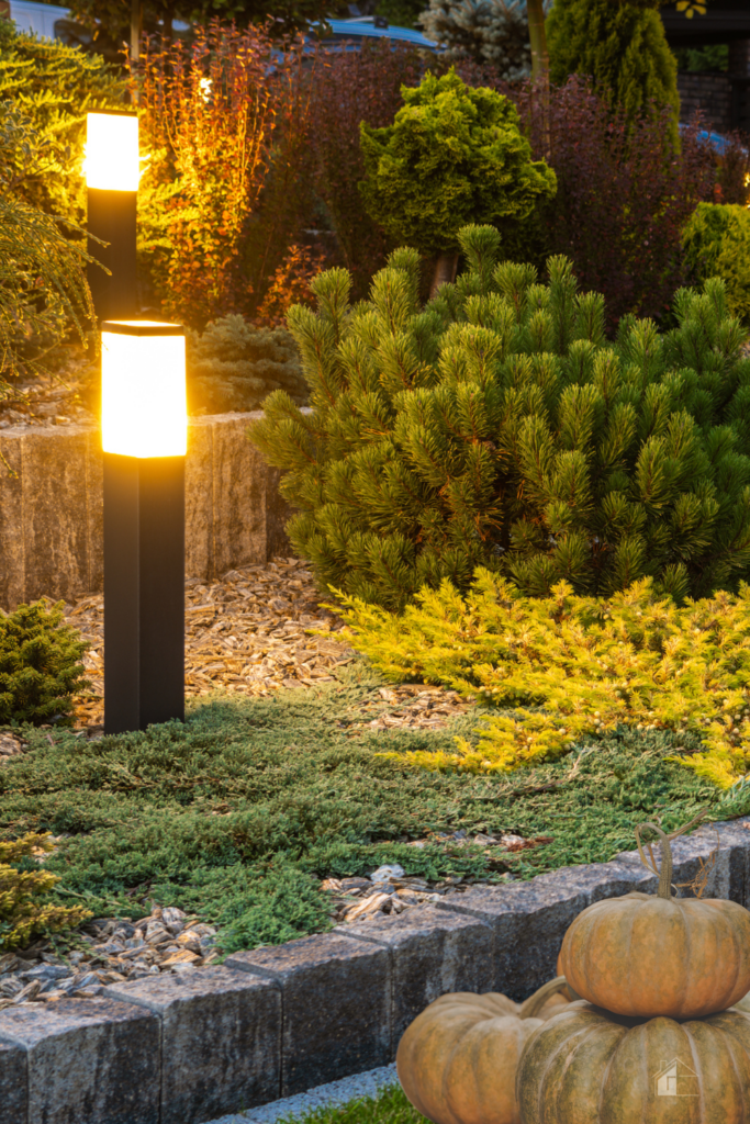 A beautifully lit fall garden with glowing pathway lights, evergreen shrubs, and decorative pumpkins, showcasing outdoor lighting ideas and seasonal decor for enhancing fall landscaping and curb appeal.