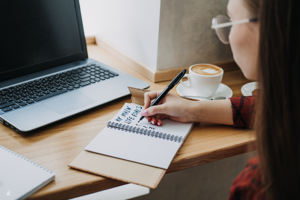 A woman writes in a goal planner at a cozy desk with a laptop and coffee, reflecting productivity, quarterly goals, and planning for success.