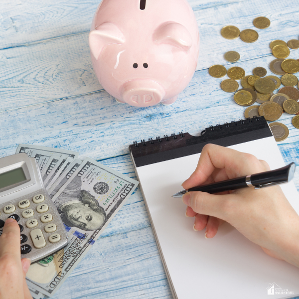 An image of a person calculating expenses. A calculator, pink piggy bank, coins, and bills are also seen in the image.