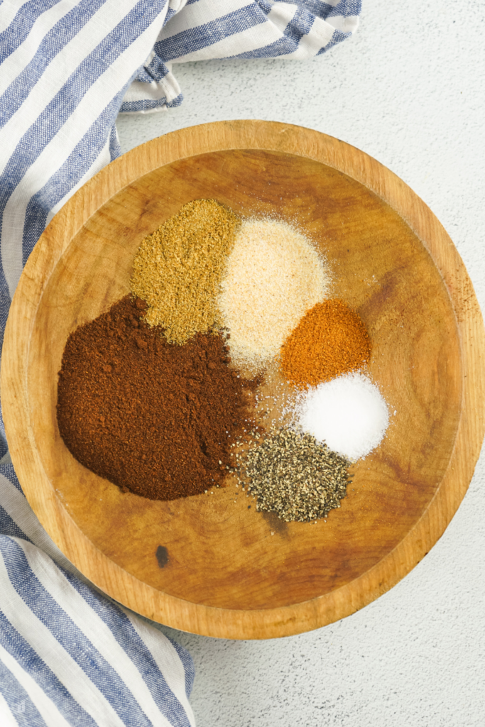 A wooden bowl filled with ingredients for an easy chili seasoning mix, including chili powder, cumin, onion powder, cayenne pepper, salt, and black pepper, ready to be blended into a homemade spice mix.