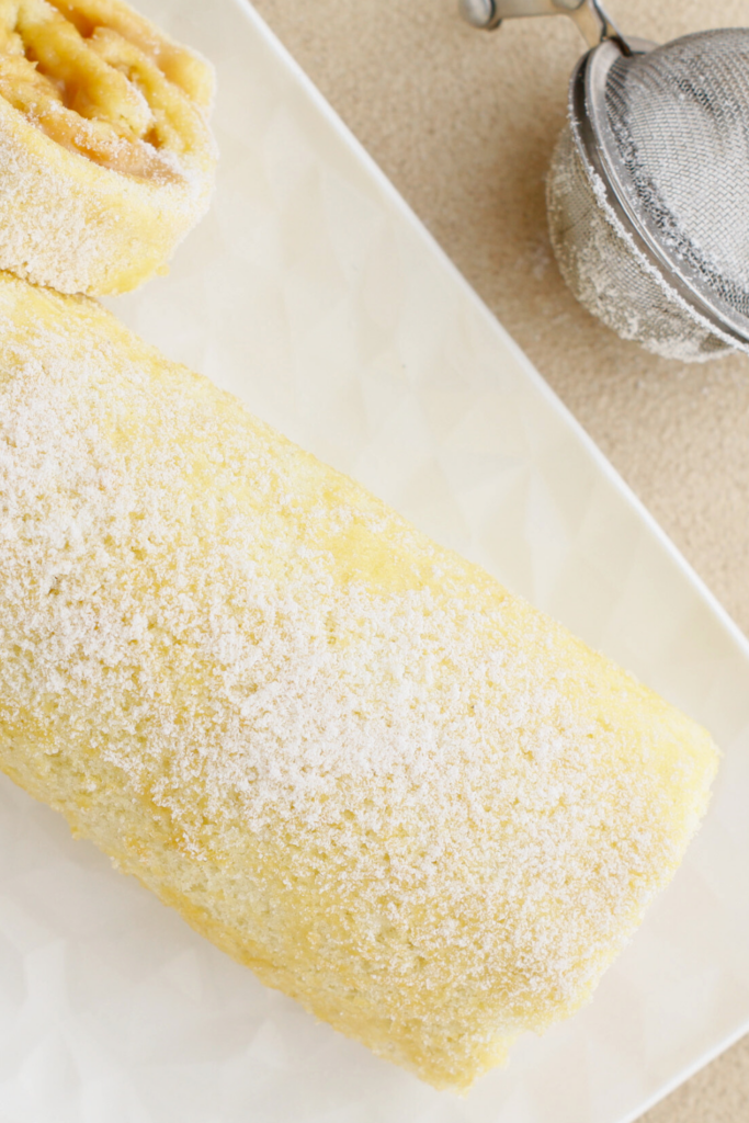 Brazo Gitano (Swiss Roll) dusted with powdered sugar, featuring its soft golden sponge and a glimpse of creamy dulce de leche filling, served on a decorative white plate.