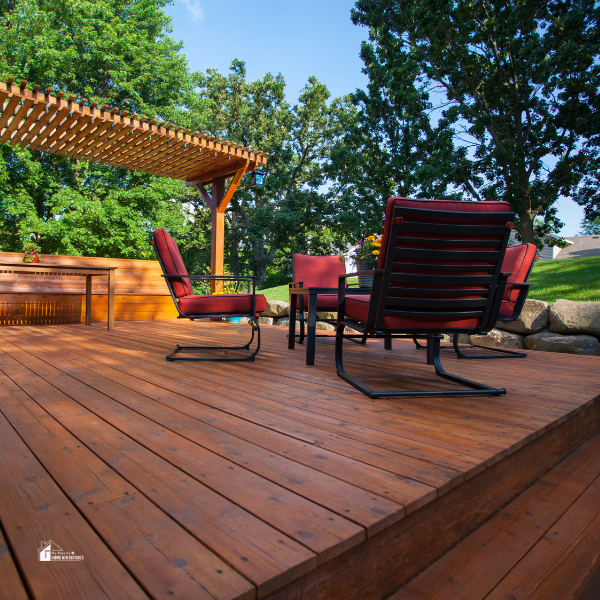 A spacious backyard deck with red chairs, a pergola, and a wooden table, set against lush green trees.