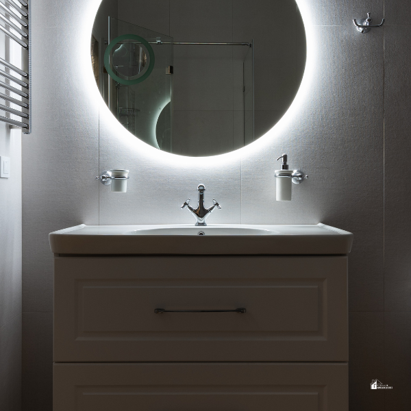 A contemporary bathroom with a circular backlit mirror above a white vanity, creating a sleek and minimalist look.