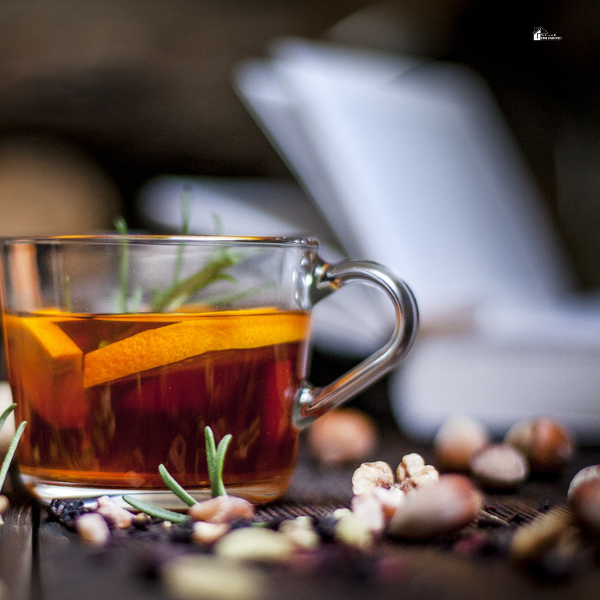 A glass cup of aromatic herbal tea garnished with orange slices and rosemary, surrounded by nuts, providing a perfect winter drink recipe to beat the chill.