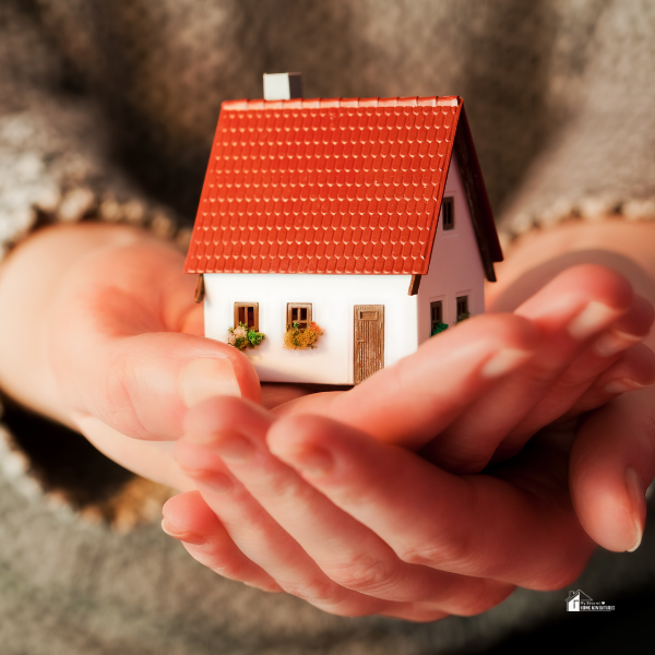 A person holding a small, detailed model house with a red roof and decorative flower boxes in their cupped hands, representing care and planning for buying a home.