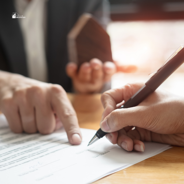 Two individuals reviewing and signing a document at a desk, with one person holding a small wooden object in the background.