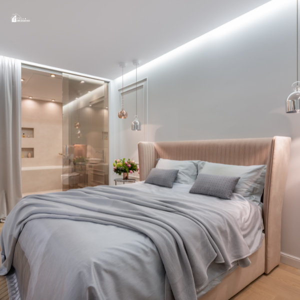 A modern bedroom with a plush upholstered bed, soft gray bedding, and hanging pendant lights, adjacent to a sleek glass-enclosed bathroom.