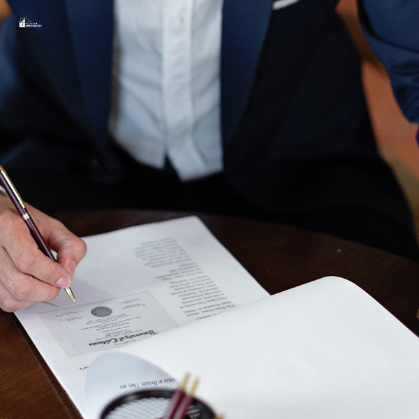 A person in a formal suit signing a certificate or legal document at a wooden table.