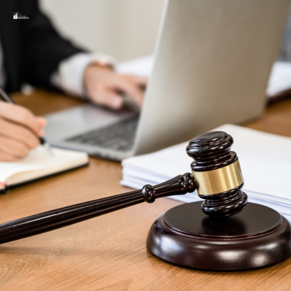 A gavel on a desk with a person working on a laptop in the background, representing legal or courtroom proceedings.