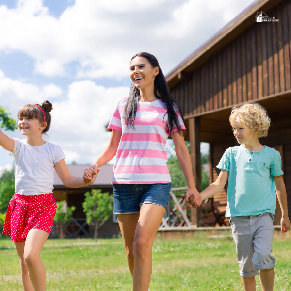 A smiling mother holding hands with two children outdoors near a cabin, representing joyful family time and outdoor adventures.