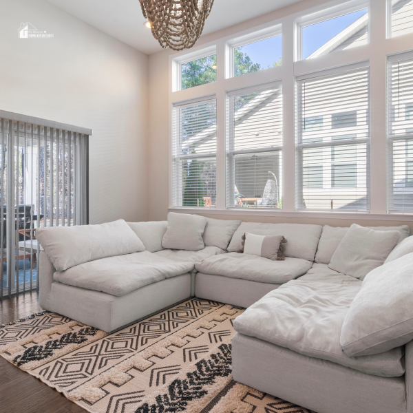 A bright modern living room featuring a large white sectional sofa and a geometric-patterned rug, with natural light streaming through tall windows.