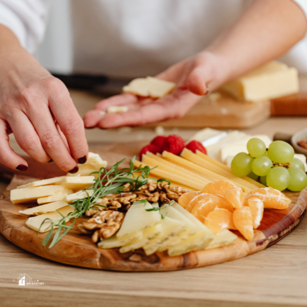 A close-up of a hand arranging cheese, nuts, fruit, and herbs on a wooden charcuterie board, showcasing ideas for French charcuterie arrangement and pairing tips.