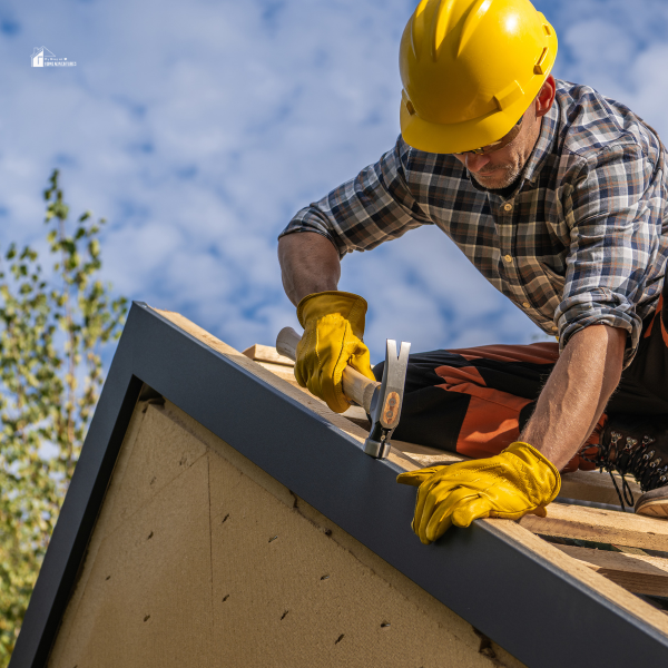 A roofer in a yellow hard hat and gloves uses a hammer while repairing the edge of a roof.