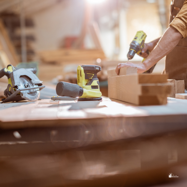 A woodworker using a drill on a workbench surrounded by tools and wooden planks in a sunlit workshop.