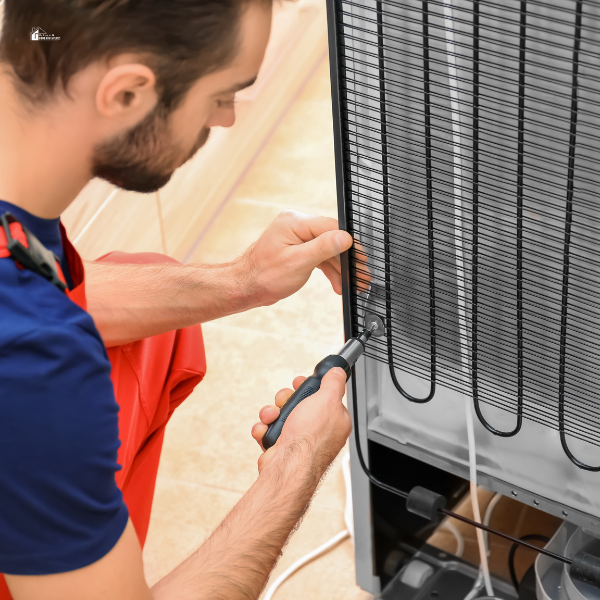 A technician in a red uniform uses a screwdriver to clean and maintain the coils on the back of a refrigerator.