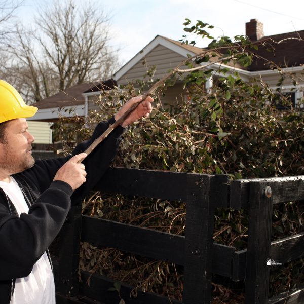 A worker wearing a yellow hard hat examines and lifts a branch from a large pile of trimmed greenery near a residential home.