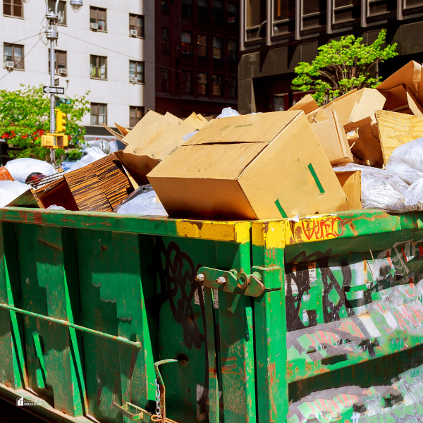 An overflowing green dumpster filled with cardboard boxes and plastic bags in an urban setting, surrounded by buildings and trees.