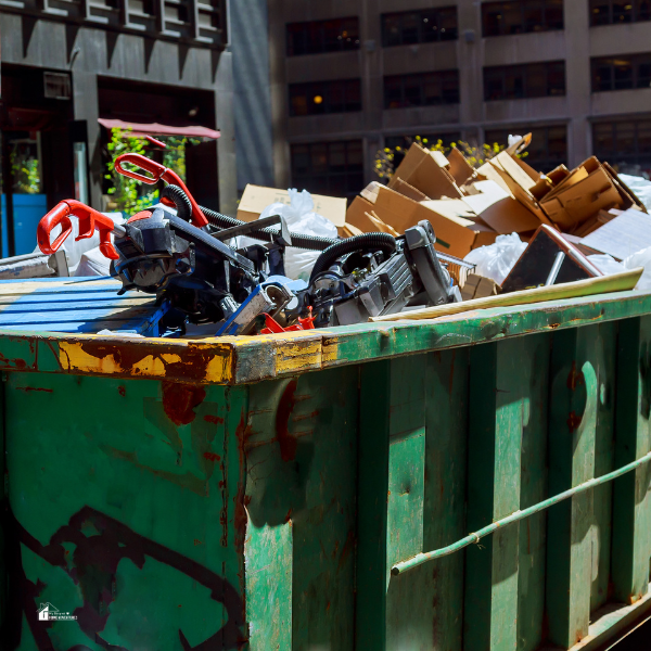 A green dumpster containing discarded cardboard, plastic, and metal debris, including vacuum cleaner parts, in a city environment.