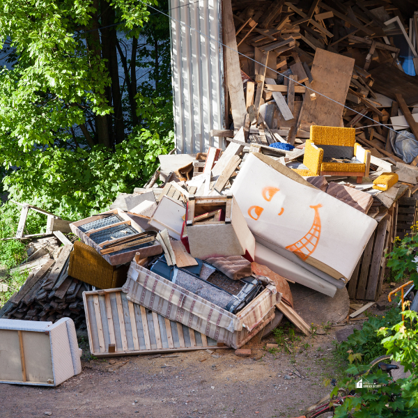 A pile of discarded furniture, mattresses, and wooden debris near a building, with greenery and a sunny outdoor setting in the background.