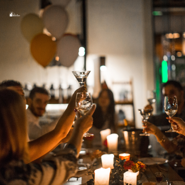 A group of people raising their glasses in a toast at a candlelit dinner party with balloons in the background.