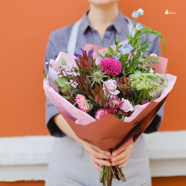 A florist holding a colorful bouquet of fresh flowers, including pink and purple blooms, wrapped in pink paper against an orange wall.