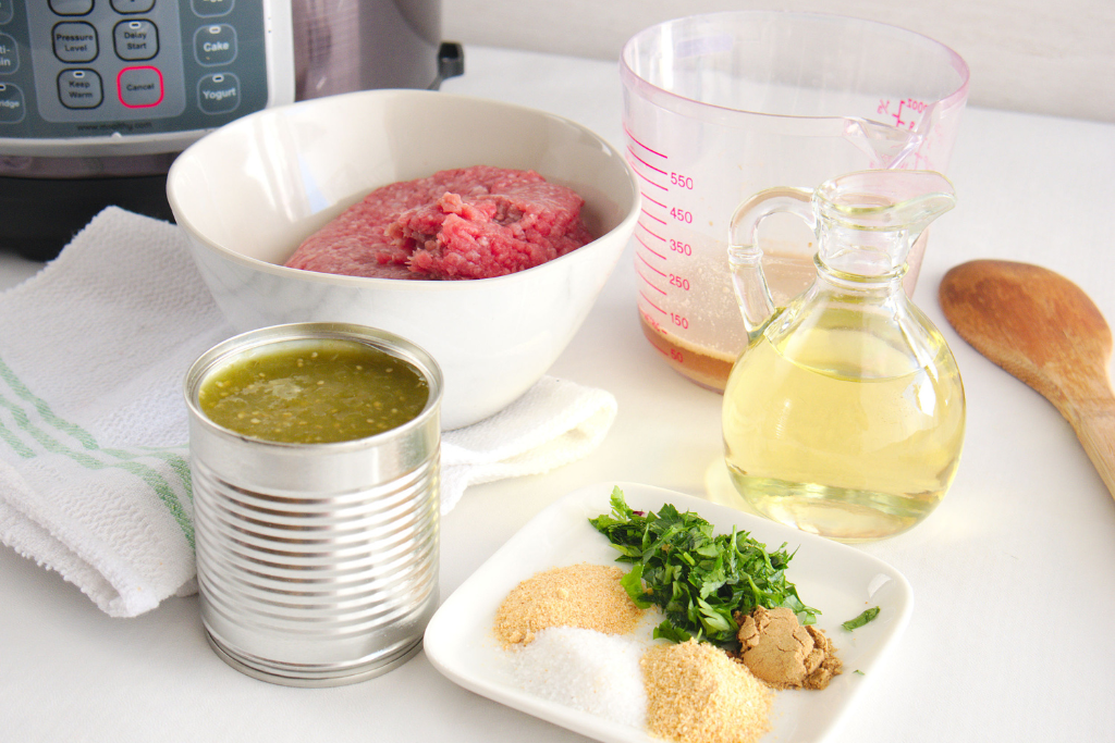 Ingredients for making pressure cooker salsa verde beef filling, including ground beef, salsa verde, broth, oil, fresh herbs, and spices, displayed with an Instant Pot in the background.