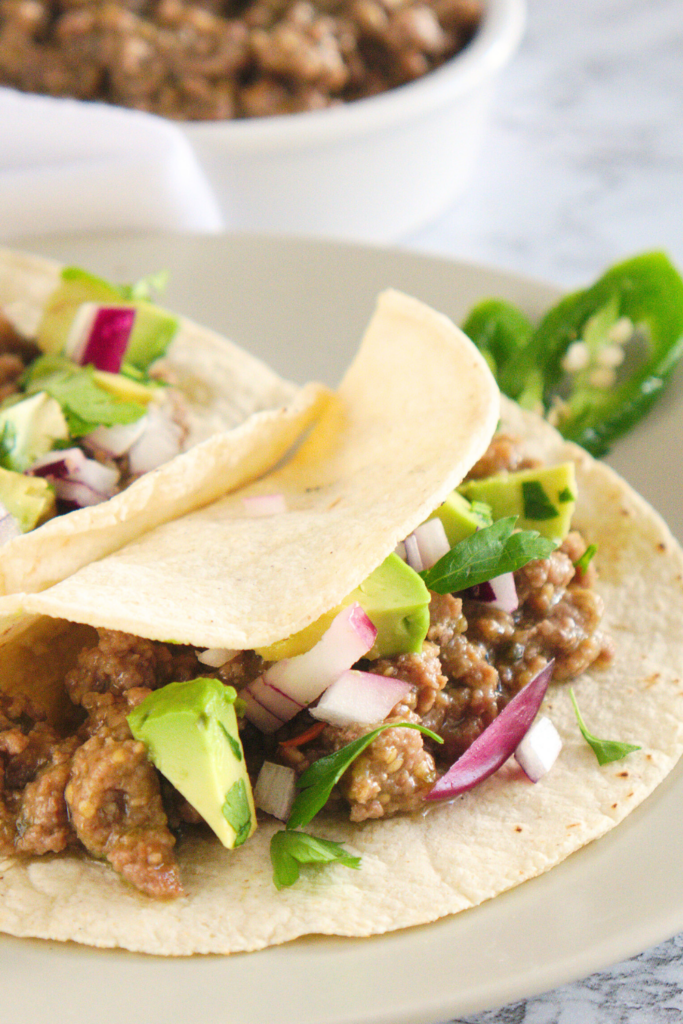 Soft corn tortillas filled with pressure cooker salsa verde beef, topped with avocado, red onion, and cilantro, served with sliced jalapeños and a bowl of Mexican beef filling in the background.