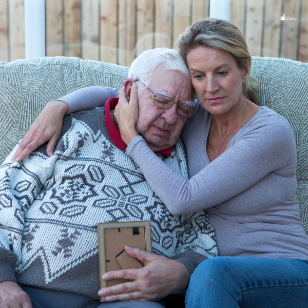 A woman comforting an elderly man on a couch, holding a picture frame in a moment of shared emotion.