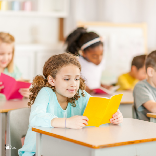 A young girl in a classroom sits at a desk reading a yellow book while other children read in the background.