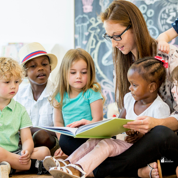 A teacher sits on the floor reading a picture book to a diverse group of young children gathered around her.