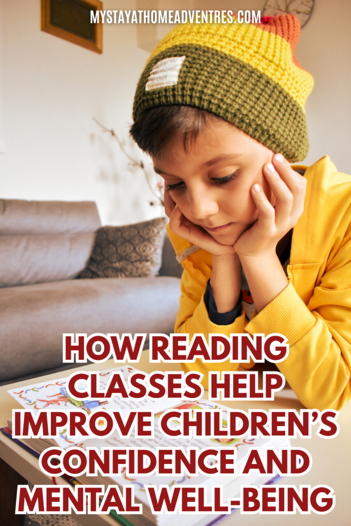 A young boy wearing a colorful knit hat sits at a table, resting his face on his hands while reading a book.